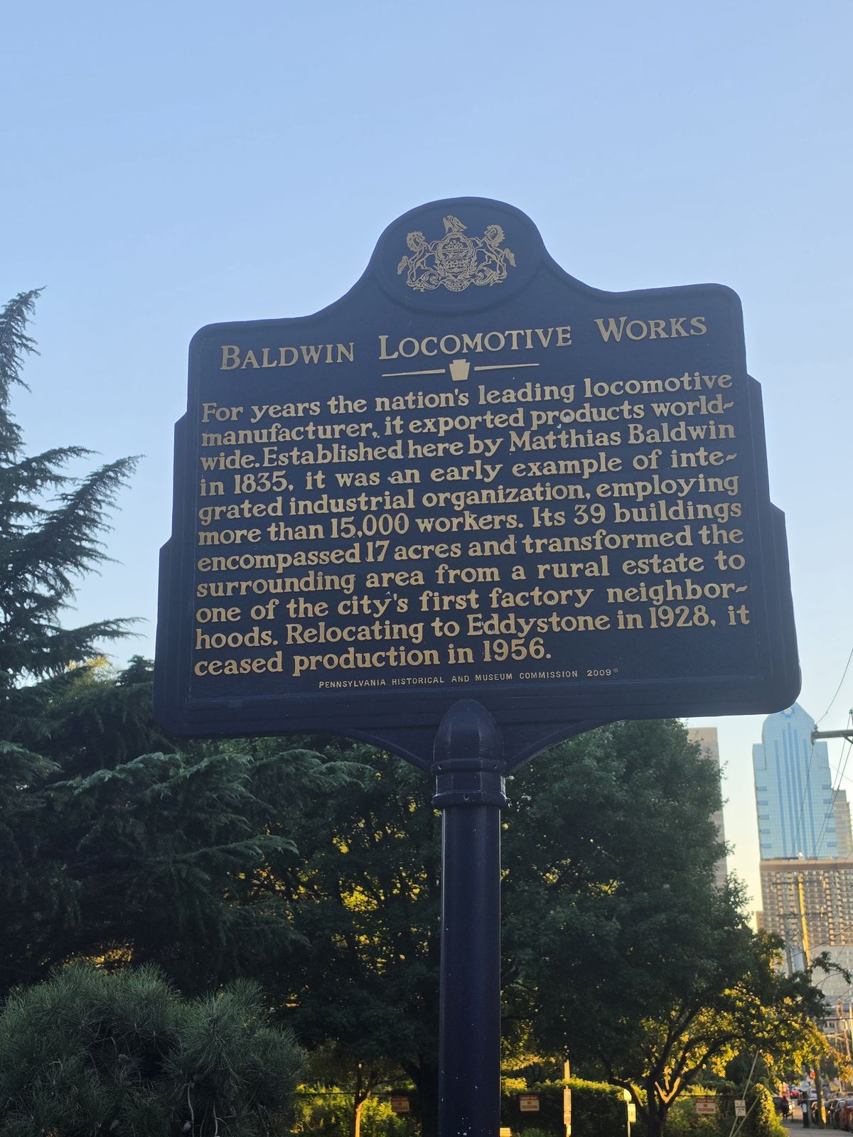 The blue Baldwin Locomotive Works historical marker at the park entrance, trees and a tower behind it.